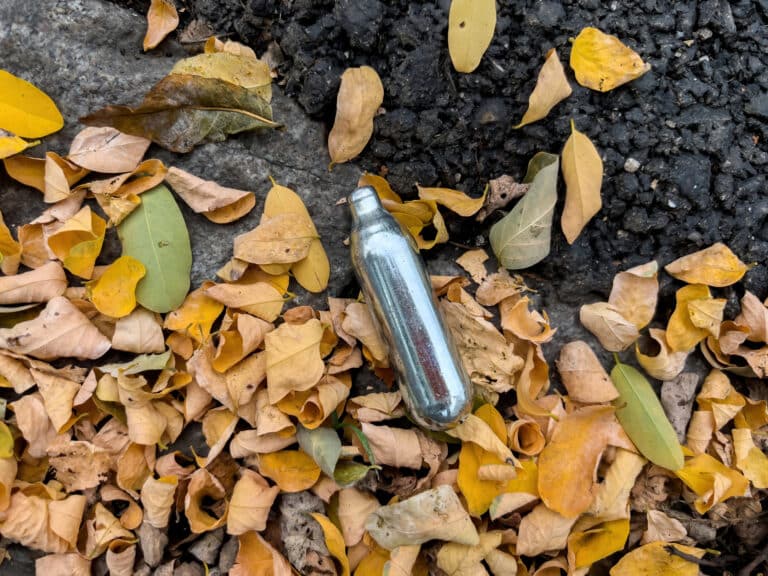 A used whipped cream charger on the ground surrounded by leaves